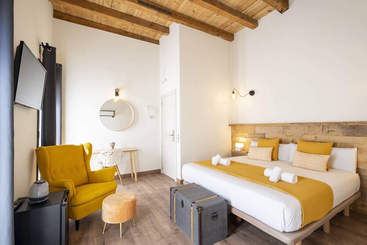 Una habitación de hotel con una cama, un sillón amarillo, una mesita, una maleta y un espejo en la pared.