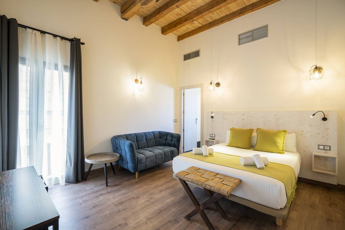 Un dormitorio con cama, mesitas de noche, una silla, una pequeña mesa y una ventana con cortinas.