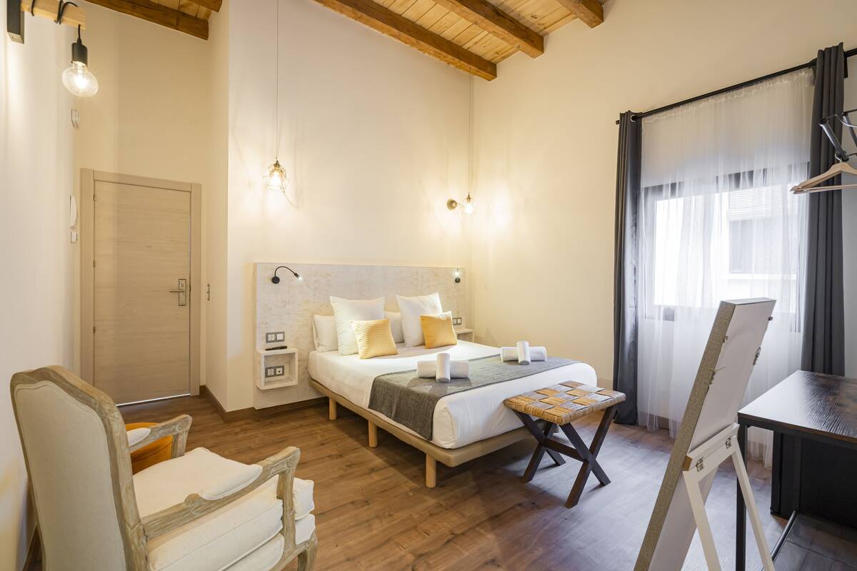 Un dormitorio con cama de madera, mesitas de noche, una silla, una pequeña mesa y un caballete con un cuadro.
