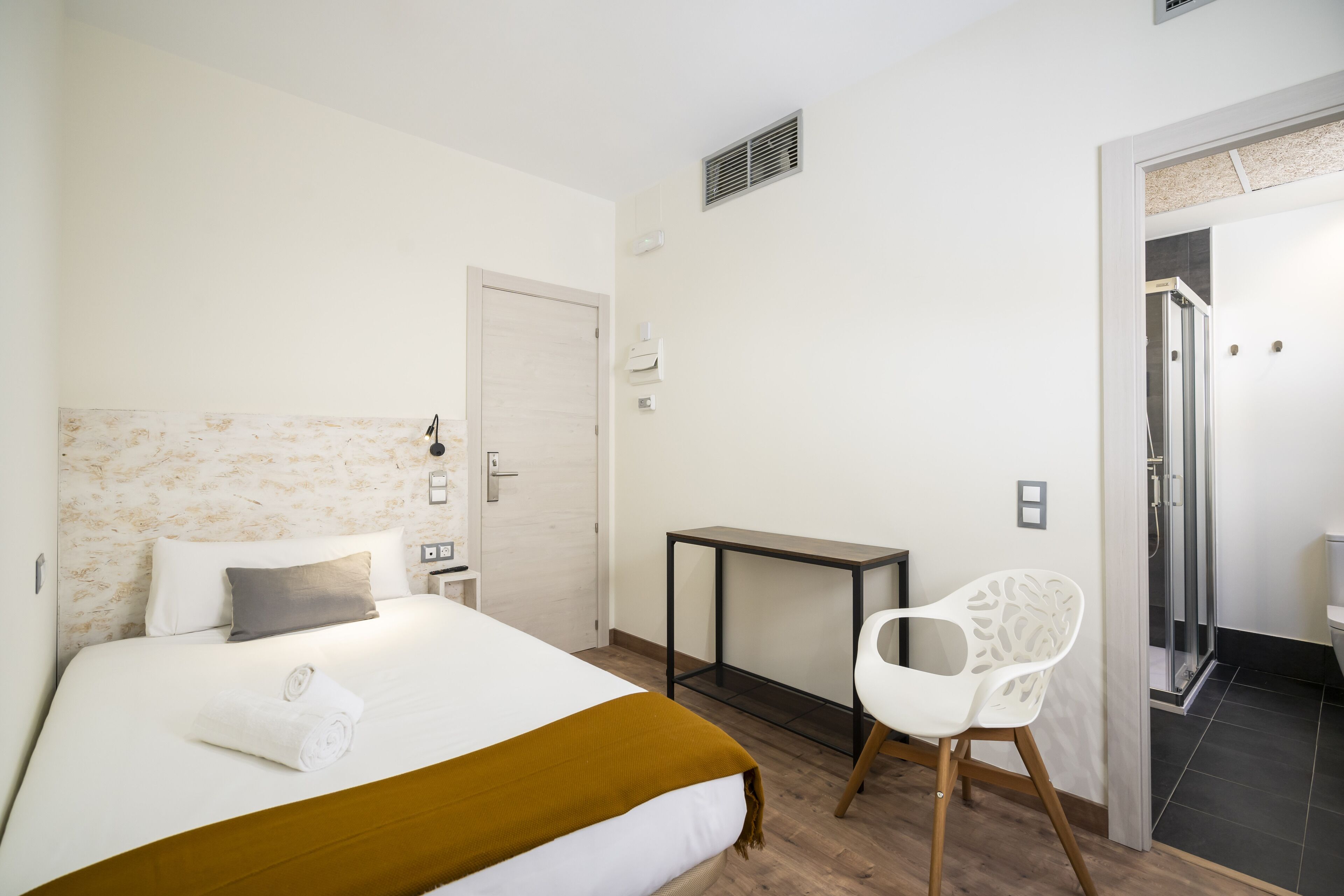 A hotel room with a bed, a desk, a chair, a door, and a shower area.