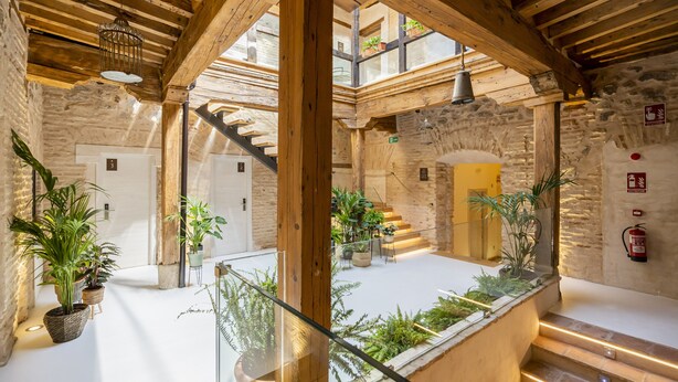 Un amplio atrio con techo alto, vigas de madera y una escalera central. Hay plantas en macetas y un extintor en las paredes.