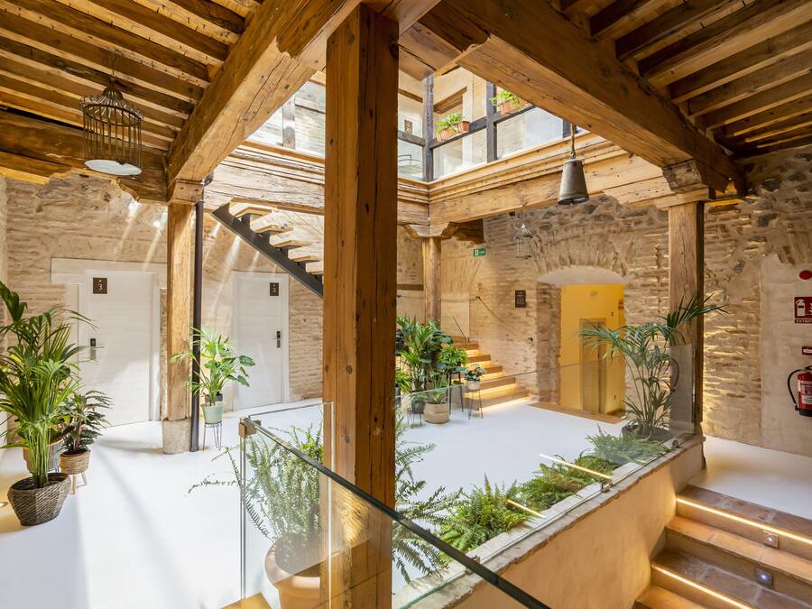 Un amplio atrio con techo alto, vigas de madera y una escalera central. Hay plantas en macetas y un extintor en las paredes.