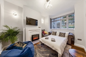 A modern bedroom with a fireplace, a sofa bed, a large window with blinds, and a potted plant. - Home Away From Home With an Indoor Fireplace! (London)
