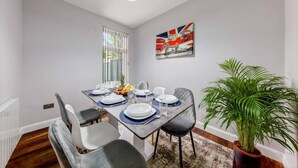Comedor con una mesa dispuesta para cuatro, incluyendo platos, vasos y un frutero. Hay una ventana con persianas, una planta en maceta y una obra de arte colgada en la pared.