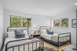 A room with two beds, each with a metal frame, white bedding, and multiple pillows. There are two windows providing a view of greenery outside.
