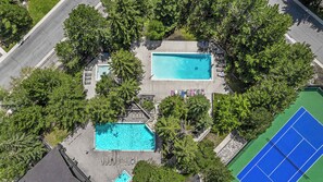 Ovenudblik over en ejendom med to swimmingpools omgivet af træer, en tennisbane og et belagt område med liggestole.