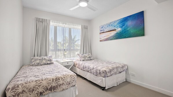 Une chambre avec deux lits, un ventilateur de plafond, une fenêtre avec des rideaux et un tableau représentant un paysage marin.