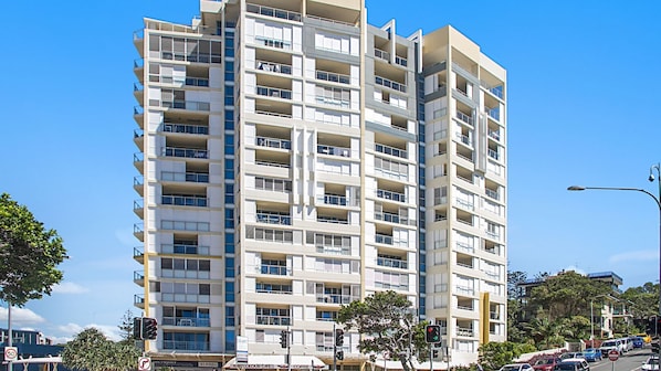 A multi-story residential building with balconies, a street in front, and traffic lights.