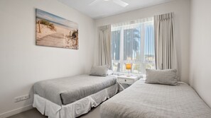 Een slaapkamer met twee bedden, een nachtkastje, een raam met gordijnen en een schilderij van een strandgezicht aan de muur.