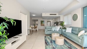 A modern living room with a flat-screen TV, a sofa, two side tables, and a dining area with chairs and a table.