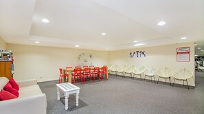 Een kamer met een witte bank, een klein tafeltje, rode en witte stoelen en een muur met een bord waarop de regels voor de fitnessruimte staan.