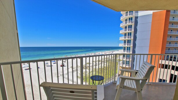 A balcony with two white chairs, a small round table, and a view of a beach with a clear blue sky. - Condo in Gulf Shores | Beachfront Views from Private Corner Balcony | Sleeps 8 (Gulf Shores)