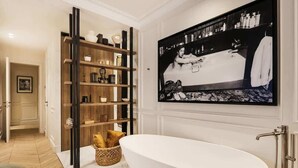 Une salle de bain moderne avec une baignoire sur pieds, une étagère en bois contenant divers objets et une grande photographie en noir et blanc accrochée au mur.