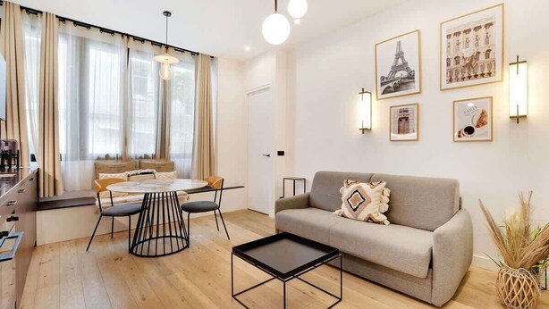 Un salon moderne comprenant un canapé, une table basse, une table à manger et des chaises. Des tableaux encadrés ornent le mur, et une fenêtre est agrémentée de rideaux.