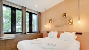 Une chambre d’hôtel moderne avec un grand lit, une tête de lit en bois et une fenêtre avec des rideaux. On y trouve des étagères murales ornées d’objets décoratifs et un luminaire suspendu.