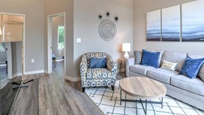 A modern living room with a patterned armchair, a grey sofa with blue cushions, a round coffee table, and a large abstract painting on the wall.