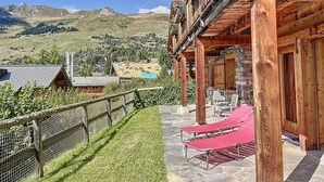 Un chalet en bois avec une façade en pierre, un balcon doté de fauteuils rouges et une vue sur un village de montagne.