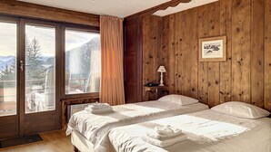 A bedroom with two beds, wooden paneling, a window with a mountain view, a framed picture on the wall, and a bedside table with a lamp. - Living room with fireplace, kitchen bar with vitroceram, Nespresso coffeemachine and dishwasher, thr (Verbier)
