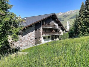 A two-story stone building with a balcony, surrounded by greenery and mountains in the background.