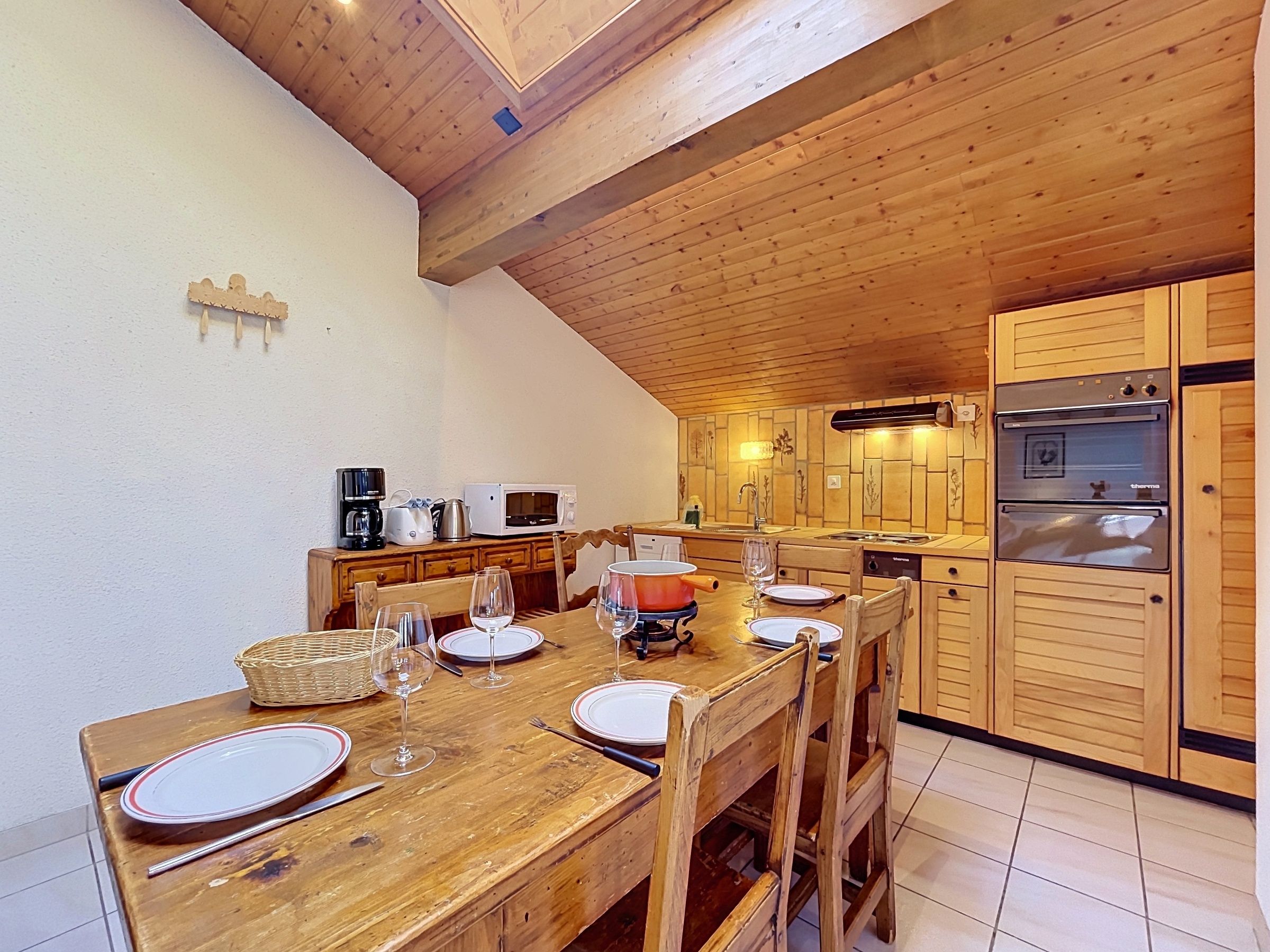 A wooden dining table set with plates, glasses, and cutlery, a microwave, and a coffee maker in a kitchen with wooden cabinets and a tiled floor.