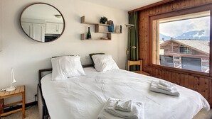 Un dormitorio con cama, mesita de noche, espejo, ventana con vista a edificios y repisa con objetos.
