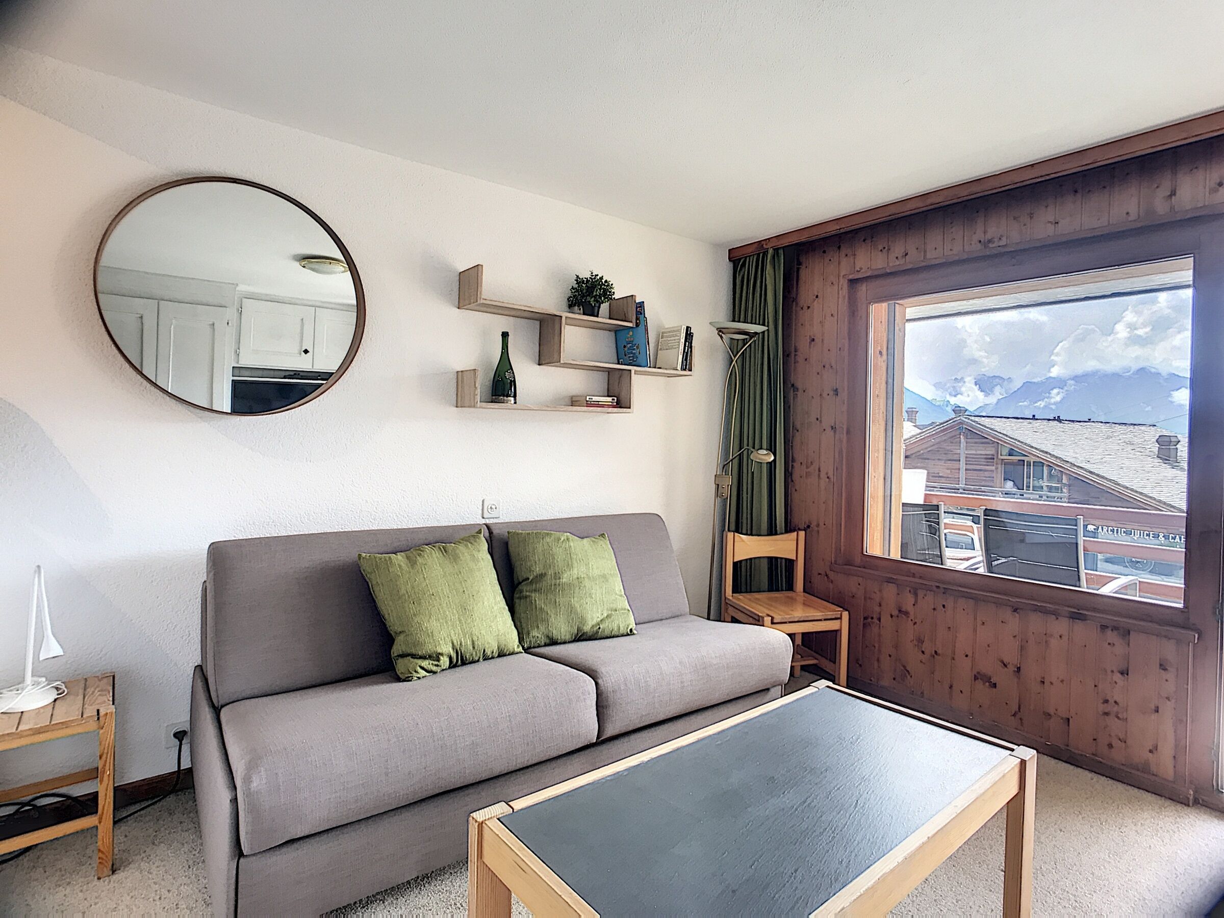 Un salon avec un canapé gris, un miroir rond, une petite table et une fenêtre donnant sur des immeubles.