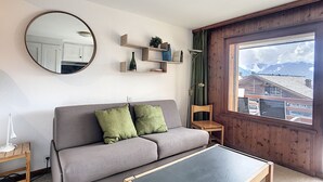 Una sala de estar con un sofá gris, un espejo redondo, una mesita y una ventana con vista a edificios.