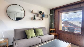 Un salon avec un canapé gris, un miroir rond, une petite table et une fenêtre donnant sur des immeubles.