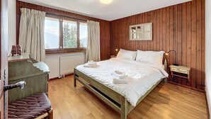 A bedroom with a wooden paneled wall, a large bed with white linens, a wooden bench, a small table with a lamp, and a window with curtains. - Living room with fireplace and eating corner,, kitchen with 3 hotplates and dishwasher, one bedroom (Verbier)