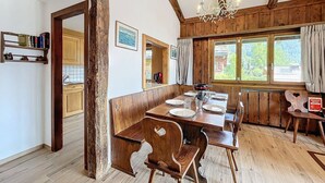 A rustic dining area with wooden furniture, a chandelier, and a view of the outdoors through large windows. - Living room with fireplace and eating corner,, kitchen with 3 hotplates and dishwasher, one bedroom (Verbier)