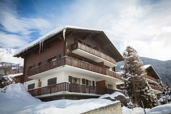 Un chalet à plusieurs étages, doté de balcons en bois, entouré de montagnes enneigées.