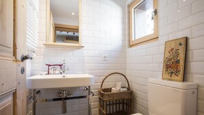Un bany amb parets revestides de rajoles blanques, un lavabo blanc, un vàter, una cistella de vimet i una obra d'art floral emmarcada a la paret.