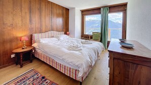 A bedroom with a wooden headboard, a bed with striped bedding, a bedside table with a lamp, a wooden dresser, and a window with a view of mountains. - Royal 6, centrally located. Close to shops and restaurants (Verbier)