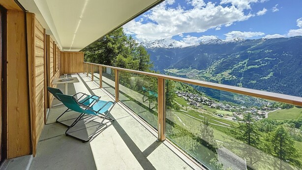 A balcony with a glass railing, a table, and a chair, overlooking a mountainous landscape with a village below. - Royal 6, centrally located. Close to shops and restaurants (Verbier)