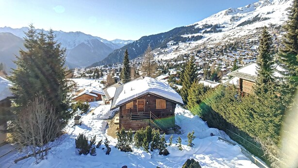 Une cabane en bois, dont le toit est recouvert de neige, entourée de neige et d’arbres, située dans une région montagneuse et offrant une vue sur des maisons et des montagnes lointaines.