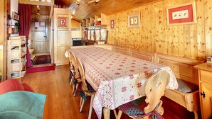 Un intérieur en bois avec une longue table à manger, des chaises, un lustre et un coin cuisine.