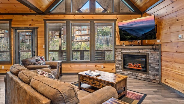 A cozy log cabin interior with a stone fireplace, leather sofas, and large windows offering views of the surrounding forest.