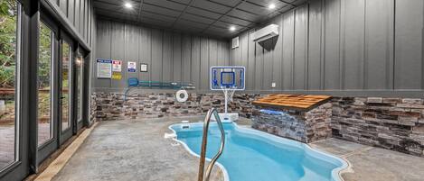 An indoor swimming pool with a basketball hoop, stone walls, and a concrete floor.