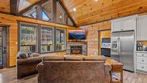 A cozy cabin interior with a fireplace, a large TV, a sofa, and a kitchen area.