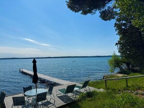 A lakeside deck with a table and chairs, a small boat, and a clear view of the water and distant shoreline.