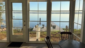 Ein Zimmer mit großen Fenstern mit Blick auf einen stillen See, eine Holzterrasse davor und ein Tisch mit Stühlen im Inneren.