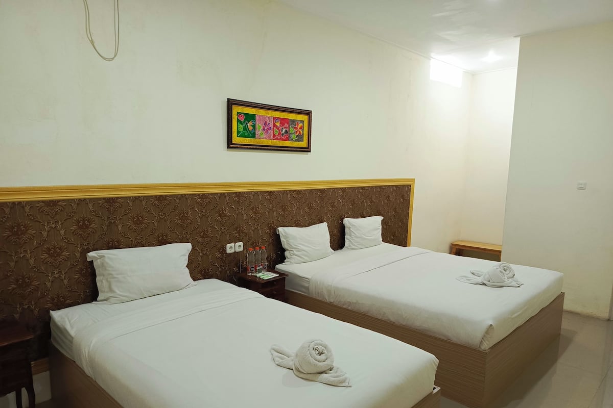 A hotel room with two beds, a wooden headboard, a small table with a bottle and a card, and a framed picture on the wall.