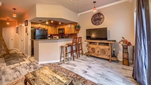 A modern kitchen with wooden cabinets, a central island, bar stools, a large TV, and a decorative vase. - SS B4 Riverside Condo (New Braunfels)