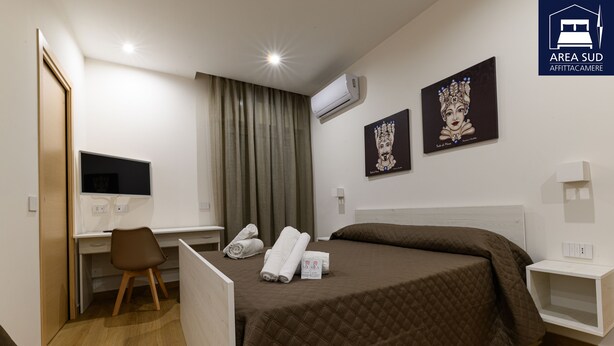 A hotel room with a bed, a desk, a chair, an air conditioner, and artwork on the walls.