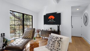 A modern living room with a large window, a sofa, a white couch, and a TV displaying the logo "HCPL Hill Country Premier Lodging". - Red Bird Cottage- Convenient 2/1 with Outdoor Space (Wimberley)