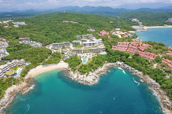 Vista aérea de un complejo turístico en la costa con varias edificaciones, una playa y un litoral rocoso.
