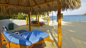 Un área de descanso frente a la playa con techo de paja, sillones de madera y vista al mar.