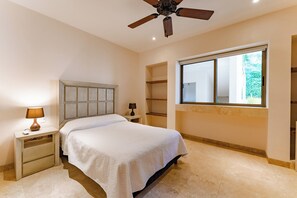 Un dormitorio con cama, mesitas de noche, ventilador de techo y ventana con vistas a la vegetación.
