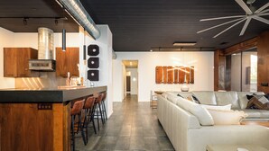 Una cocina moderna con una isla central, taburetes de bar y un horno empotrado. Se ve un espacio de sala de estar con un sofá y una mesa de centro al fondo.