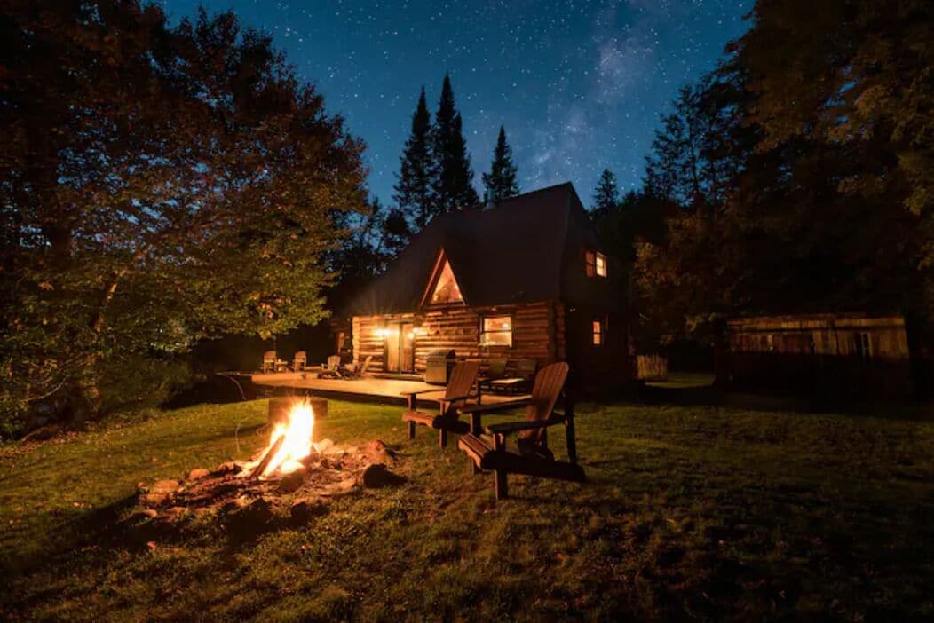Eine Blockhütte mit Feuerstelle und Sitzgelegenheit im Freien, umgeben von Bäumen und einem Sternenhimmel.